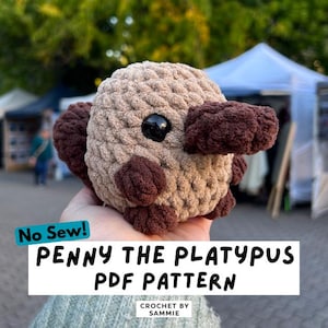 May include: A round, plush, crocheted platypus toy with a light tan body, dark brown feet, and a dark brown bill. It features a single visible black plastic eye. Text on the image reads: No Sew! PENNY THE PLATYPUS PDF PATTERN CROCHET BY SAMMIE.