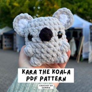 May include: A crocheted koala bear with gray fur, white inner ears, a black nose, and black eyes. The text "KARA THE KOALA PDF PATTERN" is displayed on a white rectangle. The words "CROCHET BY SAMMIE" are below the text.