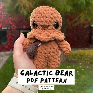 May include: A crocheted Galactic Bear plushie in shades of brown, with large black eyes and a small pouch. The text "GALACTIC BEAR PDF PATTERN" is displayed on a white rectangle. The plushie is held in a hand.