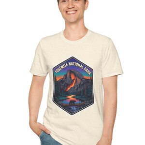 May include: A cream-colored t-shirt featuring a hexagonal graphic of Yosemite National Park. The graphic depicts a mountain, river, and trees with the text "YOSEMITE NATIONAL PARK" in a curved font. The person is wearing blue jeans.