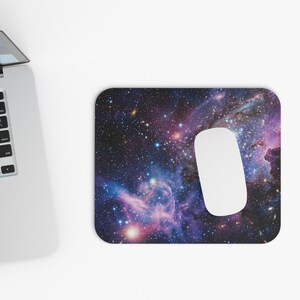 Op de afbeelding: Een rechthoekige muismat met een levendige ruimte-nevelprint in diepe paarse, blauwe en roze tinten, bezaaid met heldere witte sterren. Een gladde witte computermuis rust op het oppervlak naast een zilveren laptop met een zwart toetsenbord.