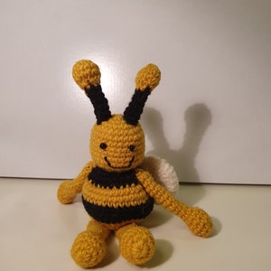 May include: A crocheted bee toy with yellow and black stripes, black eyes, and a white wing. The bee is sitting on a white surface.