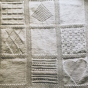 Könnte beinhalten: Eine beige gestrickte Babydecke mit verschiedenen strukturierten Quadraten. Die Decke ist mit einer Vielzahl von Maschen gearbeitet, darunter ein Stern, eine Raute, ein Zickzack und Streifen.