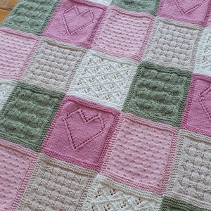 May include: A knitted baby blanket with a patchwork design featuring squares in shades of pink, green, and cream. Each square has a different stitch pattern, including hearts and cables.