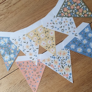 Puede incluir: Un conjunto de seis banderas de papel en forma de triángulo con motivos florales. Las banderas están decoradas con flores azules, amarillas y rosas sobre un fondo blanco. Las banderas están enhebradas juntas con una cuerda blanca.