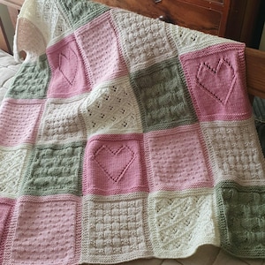Op de afbeelding: Een gebreid babydekentje met een patchwork ontwerp met vierkanten in roze, groen en wit met hartmotieven.