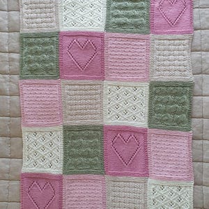 May include: A knitted baby blanket with a patchwork design featuring squares in pink, white, and green. Each square has a different stitch pattern, including hearts and diamonds.