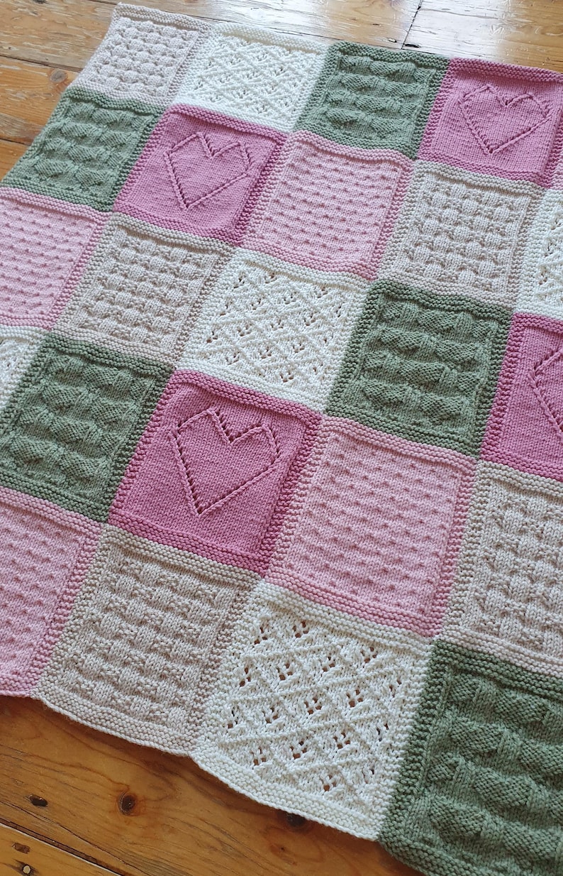 May include: A knitted baby blanket with a patchwork design featuring squares in shades of pink, green, and cream. Each square has a different stitch pattern, including hearts and cables.