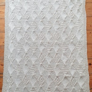 Könnte beinhalten: Eine cremefarbene gestrickte Decke mit einem Rautenmuster. Die Decke ist aus weichem Garn gefertigt und hat eine dezente Textur.