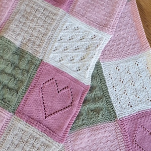 May include: A knitted baby blanket with a patchwork design featuring squares in shades of pink, green, and cream. The squares have different patterns, including hearts, cables, and lace.