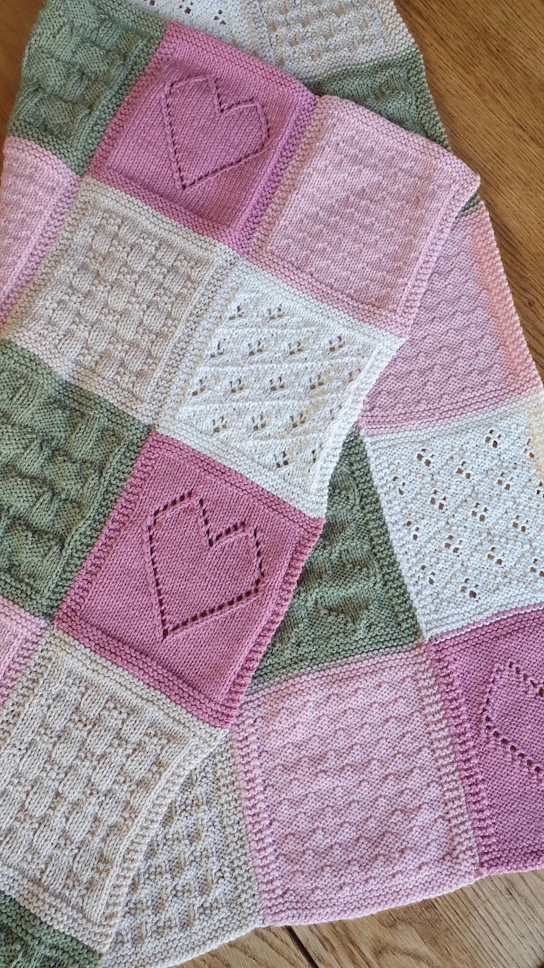 May include: A knitted baby blanket with a patchwork design featuring squares in shades of pink, green, and cream. The squares have different patterns, including hearts, cables, and lace.