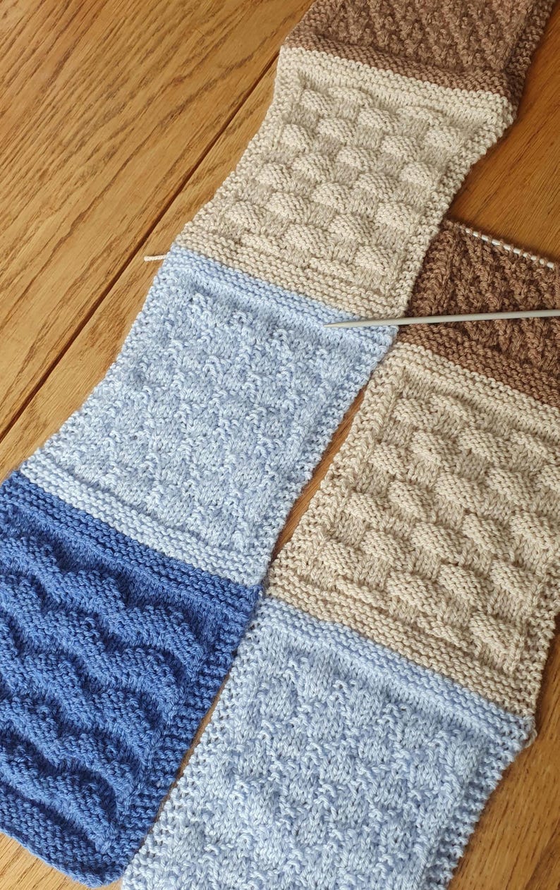May include: A knitted scarf with different colored sections. The scarf is made with a variety of stitches, including a basket weave pattern, a cable stitch, and a seed stitch. The scarf is in shades of brown, beige, light blue, and dark blue.