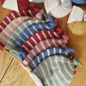 Op de afbeelding: Een verzameling gestreepte gebreide baby mutsen in verschillende kleuren, waaronder rood, roze, blauw en groen, met bijpassende bollen garen op de achtergrond. De mutsen hebben een geknoopt uiteinde en liggen op een houten oppervlak.