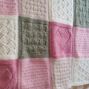 May include: A knitted baby blanket with a patchwork design featuring different patterns and colors, including pink, white, and green. The blanket is made of soft yarn and has a delicate texture.