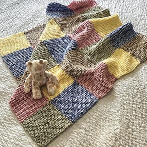 May include: A hand-knitted blanket featuring a patchwork design of square blocks in shades of yellow, blue, pink, and olive green. A small, plush giraffe toy with a brown and cream spotted pattern is positioned on top of the textured knit fabric.
