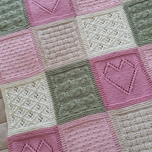 May include: A knitted baby blanket with a patchwork design featuring squares in shades of pink, green, and white. Each square has a different stitch pattern, including hearts and other geometric shapes.