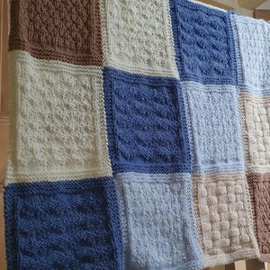 May include: A knitted baby blanket with a patchwork design in shades of brown, white, and blue. The blanket is made up of squares with different knit patterns.