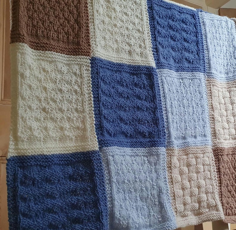 May include: A knitted baby blanket with a patchwork design in shades of brown, white, and blue. The blanket is made up of squares with different knit patterns.