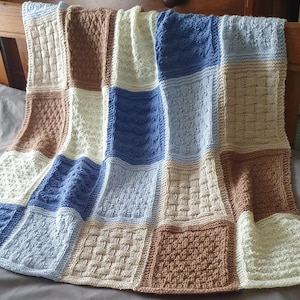 May include: A knitted baby blanket with a patchwork design in shades of brown, blue, and cream. The blanket is made up of squares with different textures and patterns.