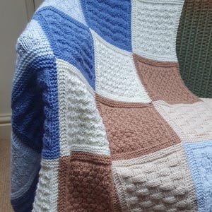May include: A knitted baby blanket with a patchwork design in shades of blue, white, and brown. The blanket is made of different textures and patterns, creating a unique and cozy look.