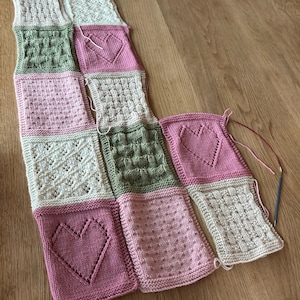 May include: A knitted blanket made of squares in pink, green, and white with different patterns, including hearts, dots, and woven designs.