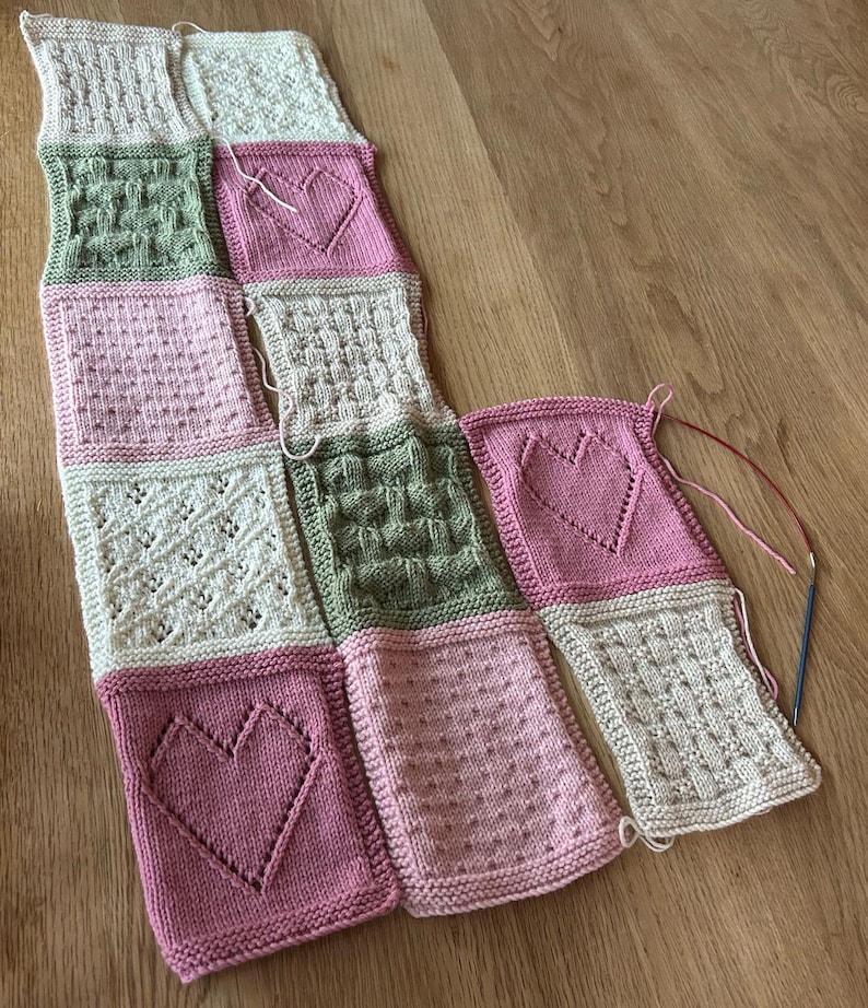 May include: A knitted blanket made of squares in pink, green, and white with different patterns, including hearts, dots, and woven designs.