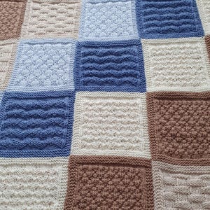 May include: A knitted baby blanket with a patchwork design in shades of brown, beige, and blue. The blanket is made up of squares with different knit patterns.