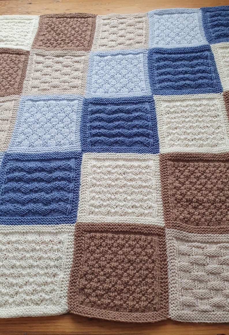 May include: A knitted baby blanket with a patchwork design in shades of brown, beige, and blue. The blanket is made up of squares with different knit patterns.