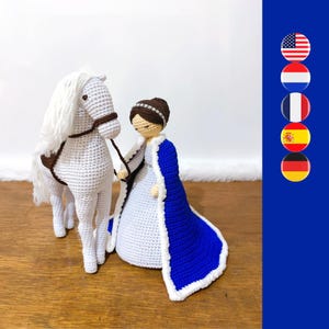 Op de afbeelding: Een wit gehaakt paard met een bruin zadel en een witte gehaakte pop die een blauwe cape en een witte jurk draagt. De pop houdt de teugels van het paard vast.
