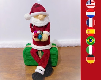 Puede incluir: Una figura de Papá Noel tejida a ganchillo con gorro y traje rojo con ribetes blancos, sosteniendo un grupo de pequeñas campanas metálicas de colores. La figura está sentada sobre una caja de regalo verde con un lazo dorado.