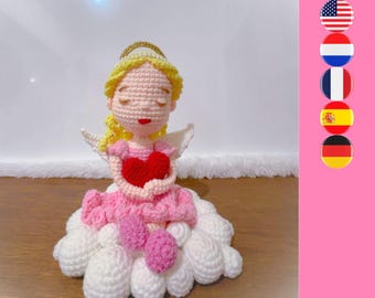 Peut inclure: Une poupée ange au crochet avec des cheveux en laine blonde, une auréole dorée et des ailes blanches. La poupée porte une robe rose, tient un cœur rouge et est assise sur un amas de nuages blancs au crochet aux yeux fermés.