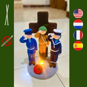 Puede incluir: Tres figuras de punto de lana se encuentran de pie frente a una cruz marrón, saludando. Las figuras están vestidas como un soldado, un marinero y un piloto. Una pequeña amapola roja se encuentra frente a una vela encendida sobre una base gris.