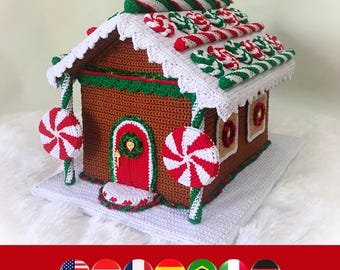 May include: A brown crocheted gingerbread house featuring a red door, white scalloped roof trim, and candy-themed decorations including red and white peppermint swirls and striped candy canes. The structure sits on a white crocheted base.