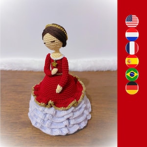 May include: A crocheted doll wearing a red dress with gold trim and a white ruffled skirt. The doll has brown hair and a crown.