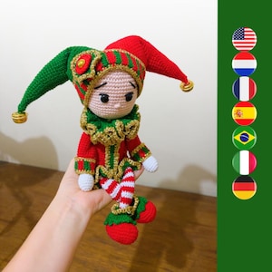 May include: A crocheted jester doll with a red and green hat, a red and green costume, and white and red striped legs. The doll is sitting on a hand.