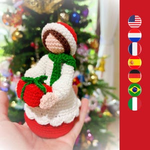 May include: A handmade crochet doll featuring a white dress with a scalloped hem, a green scarf, and a red hat. The doll has a light beige face and is holding a small, round red gift wrapped with a green yarn bow, showcasing intricate stitching.