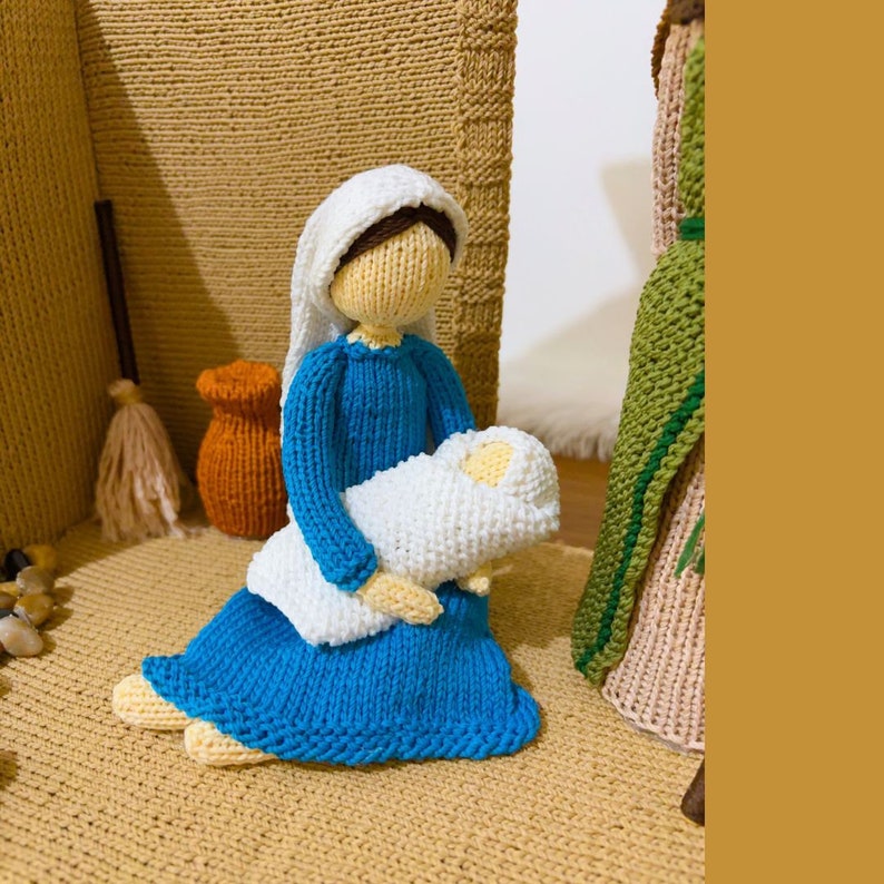 May include: Knitted figurine of Mary holding a baby Jesus, both dressed in white and blue, in a nativity scene setting.