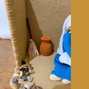 May include: A knitted figurine of a woman wearing a blue dress and a white shawl, sitting in a small, brown, woven basket. The basket is decorated with small stones and a broom. There is a small orange knitted pot in the corner of the basket.