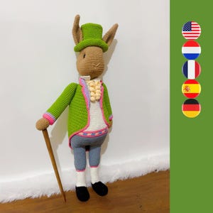 May include: A green crocheted rabbit wearing a green hat and a white shirt with a pink and purple vest. The rabbit is holding a wooden walking stick and is standing on a white surface.