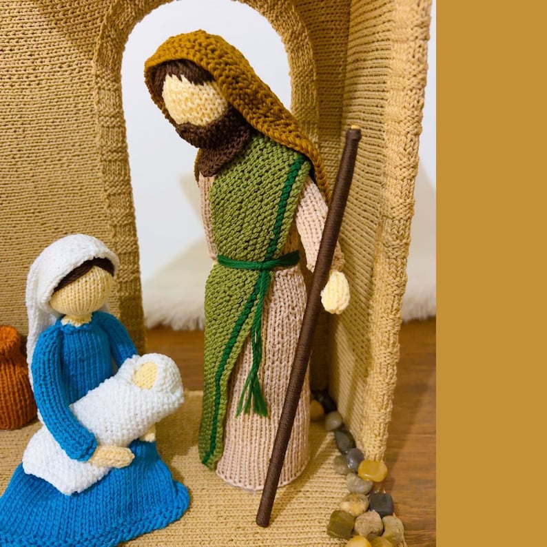 May include: Knitted nativity scene with Joseph holding a staff, Mary holding a baby, and a brown woven stable.