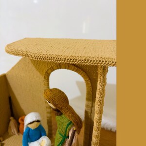 May include: A knitted nativity scene with a brown roof, a brown archway, and three figures. The figures are a woman in a blue dress, a man in a green robe, and a baby in a white blanket.