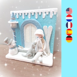 Op de afbeelding: Een gehaakt wintertafereel met een witte pop die een witte hoed draagt en een witte hond vasthoudt. De pop zit voor een blauw-wit gehaakt huis met een sneeuwpop en ski's.