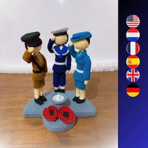 Peut inclure: Trois figurines crochetées en uniforme saluant, sur une base grise avec deux coquelicots rouges. Les figurines portent des uniformes marron, marine et bleu avec des chapeaux assortis. Une petite bougie blanche est placée entre elles.