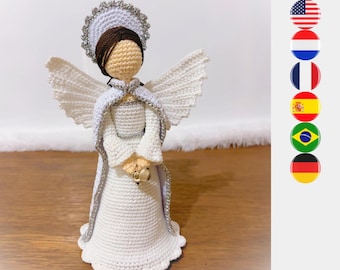 May include: A handcrafted crochet angel figurine featuring a white gown with silver trim, a matching white cape, and a halo. The figure has brown yarn hair, a beige face, and holds a small gold-colored bell in its hands, standing upright.