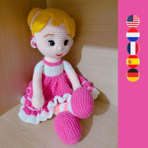 Peut inclure: Une poupée en crochet aux cheveux blonds, portant une robe rose avec des bordures blanches et des chaussures roses. La poupée est assise sur une surface en bois.