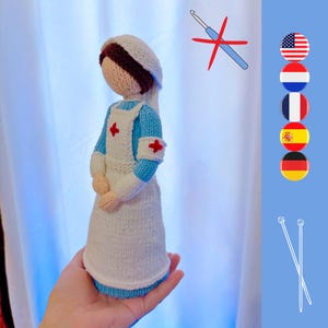 May include: A handmade knitted doll dressed as a nurse, featuring a white bonnet with a brown band, a light blue uniform with a white apron, and a red cross emblem. The doll is held in a hand.