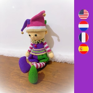 Puede incluir: Un muñeco de bufón de crochet con un traje morado y verde, un sombrero rosa y blanco con cascabeles y una cara amarilla. El muñeco está sentado sobre una superficie de madera.