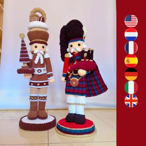 May include: Two crocheted nutcracker figures standing on circular bases. The left figure is brown and white, holding a small tree. The right figure wears a tall black hat, a red and blue plaid kilt, and carries a red bagpipe instrument.
