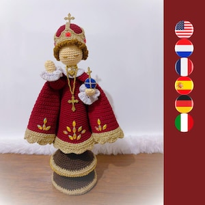Op de afbeelding: Een gehaakte pop van het Kindje Jezus van Praag, gekleed in een donkerrode mantel met gouden rand, een bijpassende kroon en witte kanten manchetten. De figuur houdt een blauwe en gouden rijksappel vast, draagt een gouden kruisketting en iriserende edelstenen.