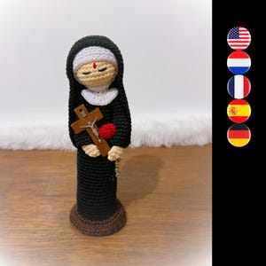 Puede incluir: Muñeca de monja de ganchillo con hábito negro, velo blanco y cruz de madera. La muñeca sostiene una rosa roja y tiene una base marrón.